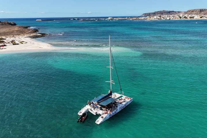 Catamaran sailing on turquoise water near a sandy beach and distant town.
