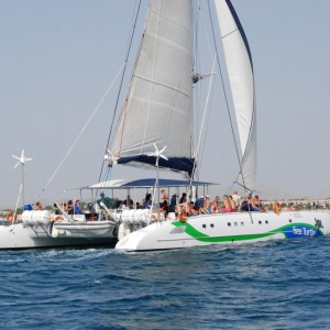 Sailing catamaran with passengers on open water near a sandy shoreline.