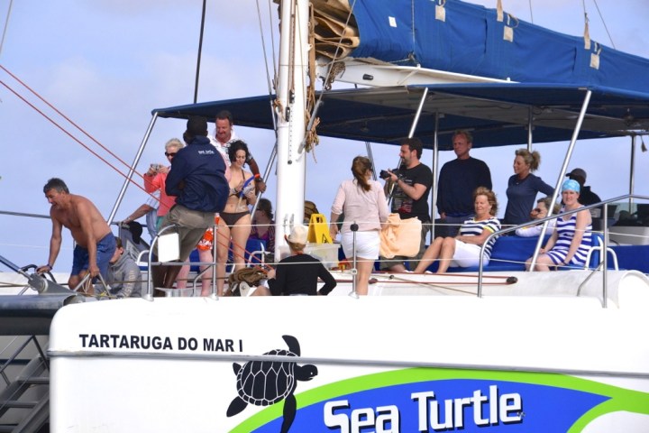 Group of people on a boat deck, some standing, some seated, with a turtle logo visible.