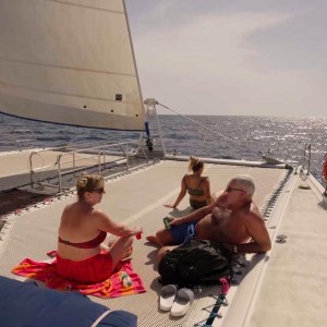 Three people relaxing on a sailboat deck under the sun near the ocean.
