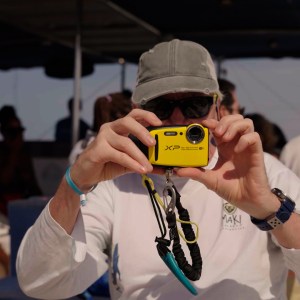 Person in hat taking a photo with a yellow camera on a boat with others in the background.