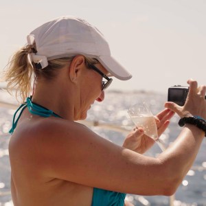 Woman on a boat taking a photo with a camera, holding a drink, and wearing a white cap and sunglasses.