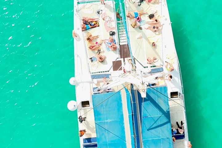 Aerial view of people sunbathing on a catamaran in turquoise water, with swimmers nearby.