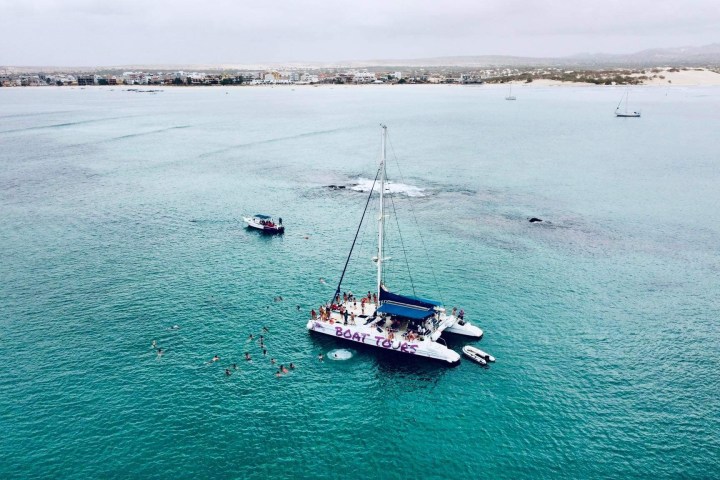 Aerial view of a sailboat labeled 'Boat Tours' on clear turquoise waters with swimmers nearby, coast in background.