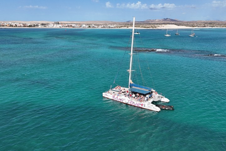 Catamaran with people on turquoise sea, distant coastline and hills.