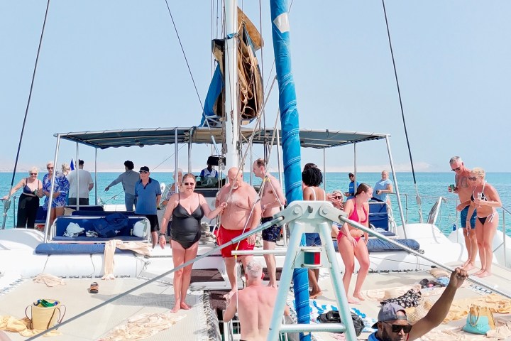 People on a catamaran enjoying sunny weather with some swimming in turquoise water.